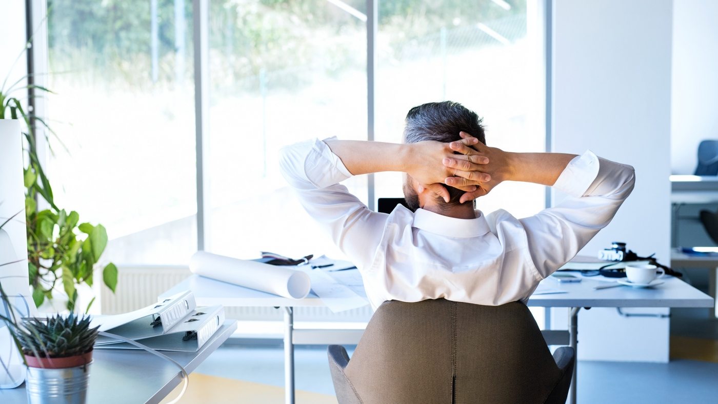 Handsome young businessman in his office, sitting at the desk, stretching arms. Rear view.
